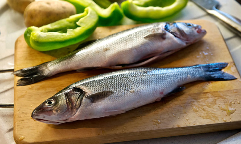 Dois peixes da espécie robalo sobre uma tábua de madeira, com pimentões e batatas em volta.